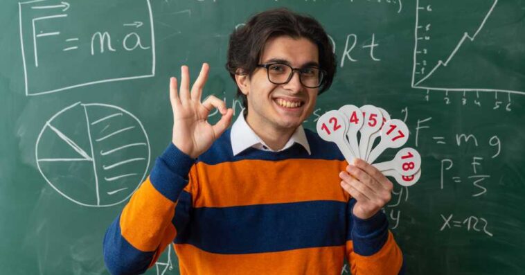 smiling young geometry teacher wearing glasses standing front chalkboard classroom holding number fans looking front doing ok sign 2 1