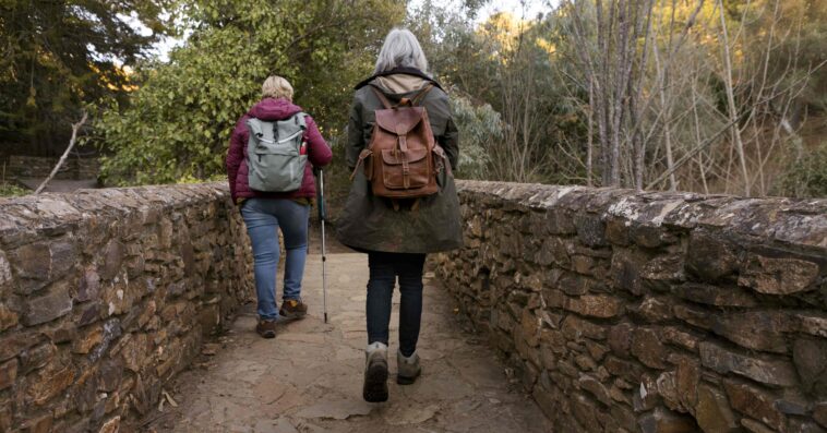 two senior women crossing stone bridge while out nature 1 1