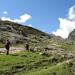 Two trekkers make their way up a lush green trail surrounded by rocky hills and blue skies, capturing the serene beauty of a trek in Kashmir.