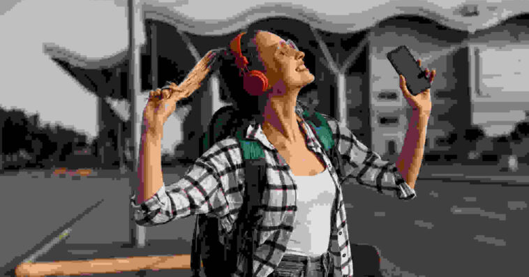 Happy traveler with backpack enjoying music on headphones while holding phone at a transport terminal